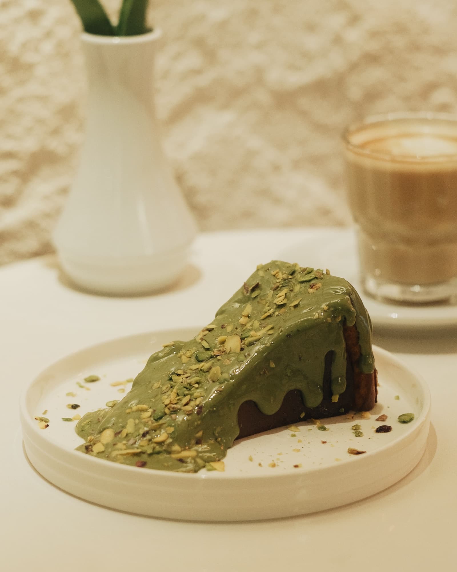 Üstü fıstık ezmesi ve kırık fıstıkla kaplı bir dilim San Sebastian; arkada bir bardak latte.
