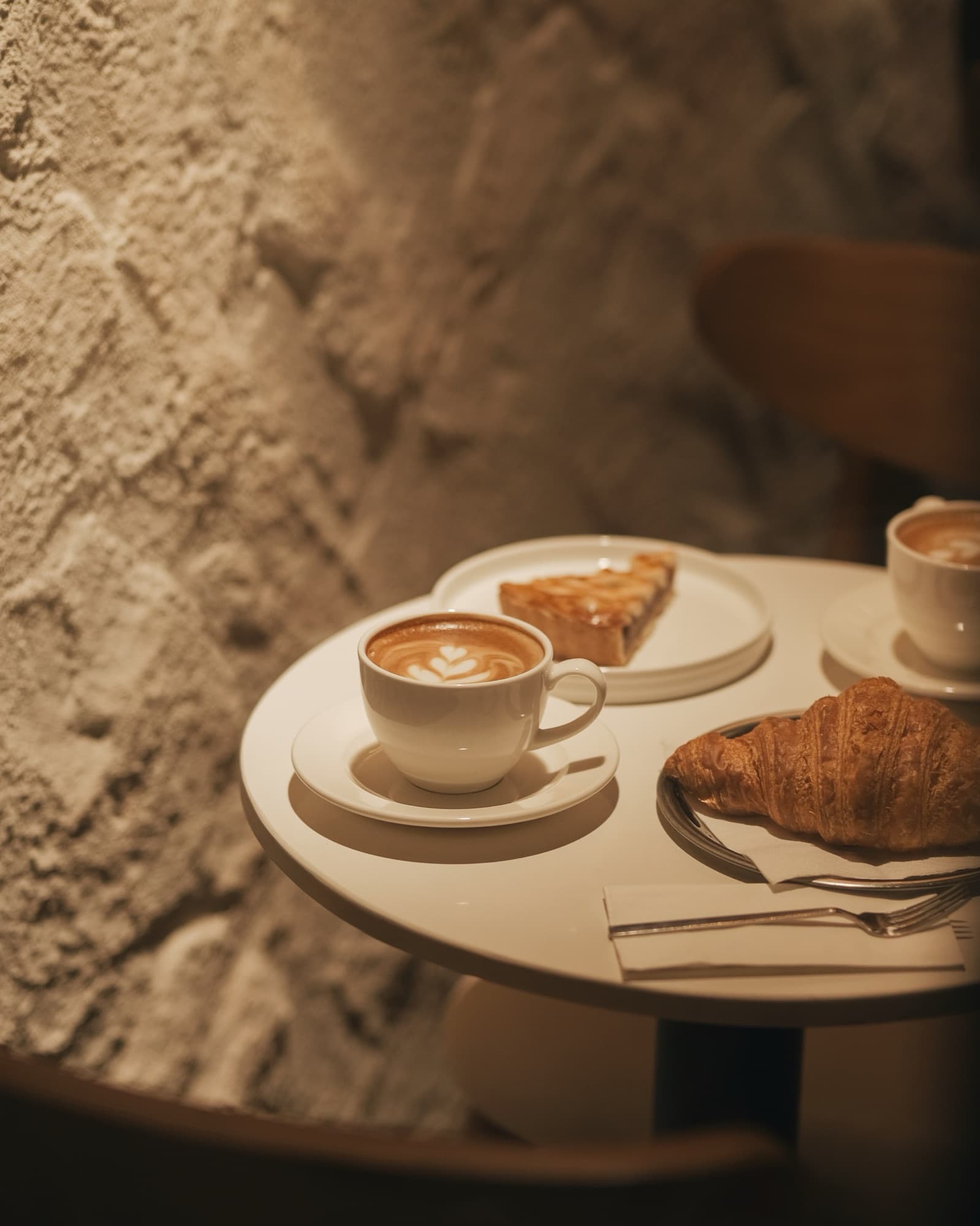 Krem sıvalı duvarın önünde yuvarlak beyaz masa: latte art'lı bir fincan, kruvasan ve bir dilim tatlı.