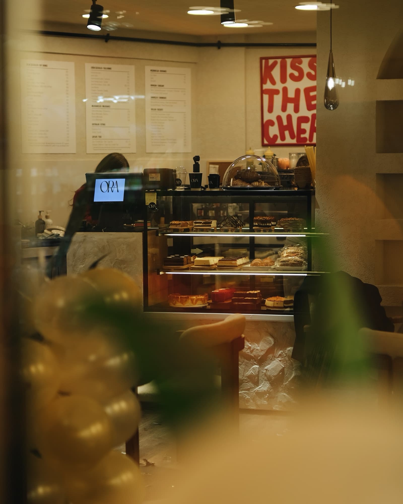 Camdan görünen ORA tezgâhı: pasta vitrini, iki dilli menü panoları ve duvarda kırmızı “Kiss the Chef” afişi.